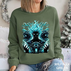 Op de afbeelding: Militairgroen sweatshirt met een opvallende afbeelding van een figuur met blauwe vlammen als haar, omringd door drie leeuwachtige wezens met felblauwe ogen. Het ontwerp is gecentreerd op de voorkant van de trui.