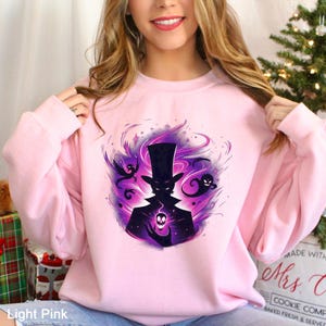 Op de afbeelding: Lichtroze sweatshirt met een afbeelding van een silhouet met een hoge hoed, die een gloeiende schedel vasthoudt. Het ontwerp bevat paarse en zwarte wervelende elementen en spookachtige figuren. De tekst "Light Pink" is linksonder zichtbaar.