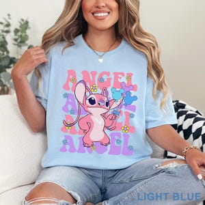 May include: Light blue t-shirt featuring a pink cartoon character holding balloons, with the word "ANGEL" repeated in a retro font. The design includes flowers in pink, purple, and yellow.