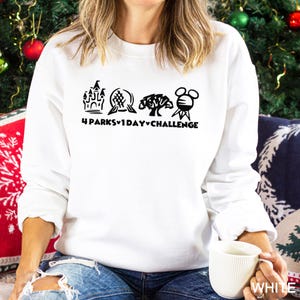 May include: White sweatshirt with a black graphic featuring four theme park icons and the text "4 Parks 1 Day Challenge." The sweatshirt is a crew neck style and is being held by a person.