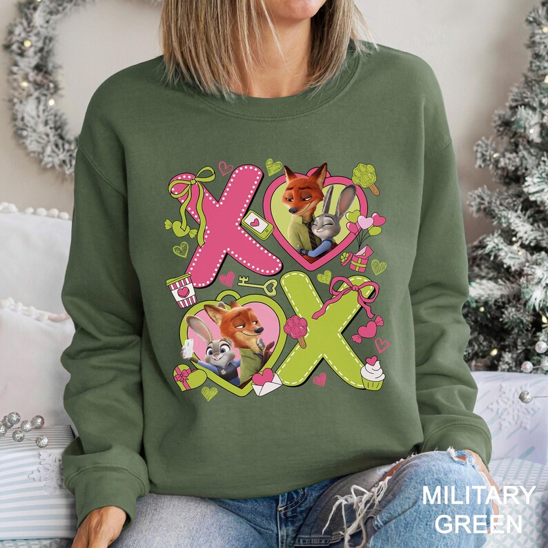 Peut inclure: Sweat-shirt vert militaire avec un motif graphique des personnages Nick Wilde et Judy Hopps de Zootopie. Le motif comprend des c&oelig;urs roses et verts et les lettres "X" et "O".