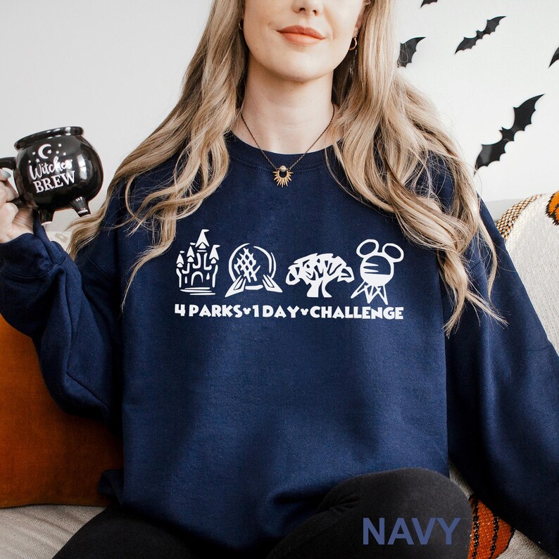 May include: Navy blue sweatshirt with a white graphic featuring four park icons and the text "4 Parks 1 Day Challenge." The word "NAVY" is printed at the bottom. A black mug with the words "Witches Brew" is held in the left hand.