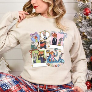 May include: Sand-colored sweatshirt featuring a collage of Zootopia characters in various poses. The design includes images of Nick Wilde, Judy Hopps, and other characters in a cartoon style. The word "Zootopia" is also visible.