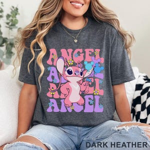 May include: Dark heather gray t-shirt featuring a pink cartoon character holding blue flowers, with the word "ANGEL" in pink and purple, and small flower graphics. The text "DARK HEATHER" is at the bottom.