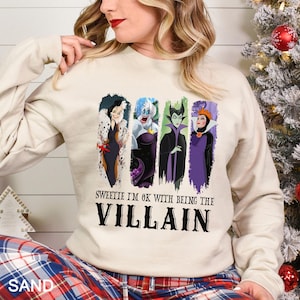 May include: A sand-colored sweatshirt featuring four cartoon villain characters in vertical panels. The text "Sweetie I'm OK with being the Villain" is printed below the characters in a bold, black font. The person is wearing plaid pajama pants.