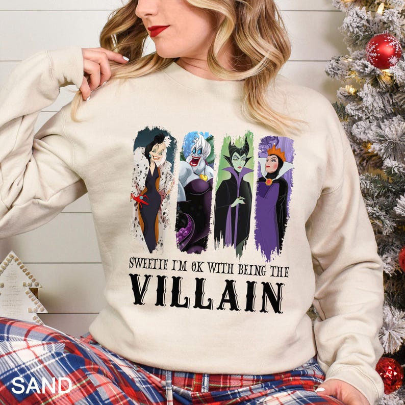 May include: A sand-colored sweatshirt featuring four cartoon villain characters in vertical panels. The text "Sweetie I'm OK with being the Villain" is printed below the characters in a bold, black font. The person is wearing plaid pajama pants.