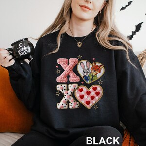 May include: Black sweatshirt featuring the letters "XO" in pink and white, with heart and star accents. The design includes a heart with cartoon characters and a heart filled with red hearts. The word "BLACK" is at the bottom.