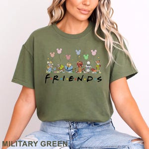 Puede incluir: Camiseta verde militar con un dise&ntilde;o de dibujos animados de personajes sosteniendo globos. La palabra "FRIENDS" est&aacute; impresa en una fuente colorida debajo de los personajes. La camiseta es de un estilo c&oacute;modo e informal.