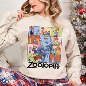 May include: A sand-colored sweatshirt featuring a colorful graphic of characters from the movie "Zootopia." The graphic is a grid of images with the word "ZOOTOPIA" in large black letters at the bottom. The sweatshirt has long sleeves.