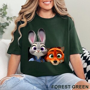 May include: Forest green t-shirt featuring a graphic of Judy Hopps and Nick Wilde from Zootopia. The cartoon characters are in a rectangular design. The text "FOREST GREEN" is printed at the bottom of the shirt.
