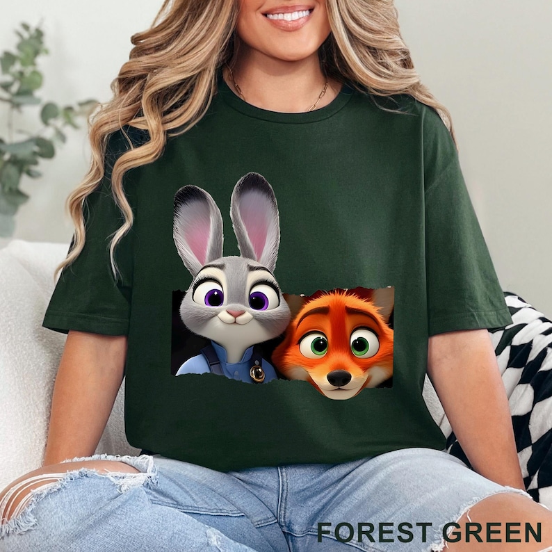 May include: Forest green t-shirt featuring a graphic of Judy Hopps and Nick Wilde from Zootopia. The cartoon characters are in a rectangular design. The text "FOREST GREEN" is printed at the bottom of the shirt.