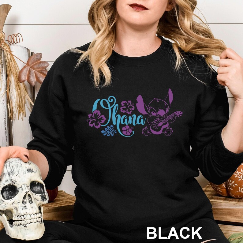 May include: Black sweatshirt featuring the word "Ohana" in blue and purple script, with a cartoon character playing a guitar. The word "BLACK" is printed at the bottom of the sweatshirt.