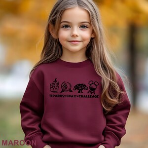 May include: A maroon sweatshirt with the text "4 Parks 1 Day Challenge" and four graphic icons. The sweatshirt is worn by a young girl with long brown hair. The word "MAROON" is printed at the bottom of the image.