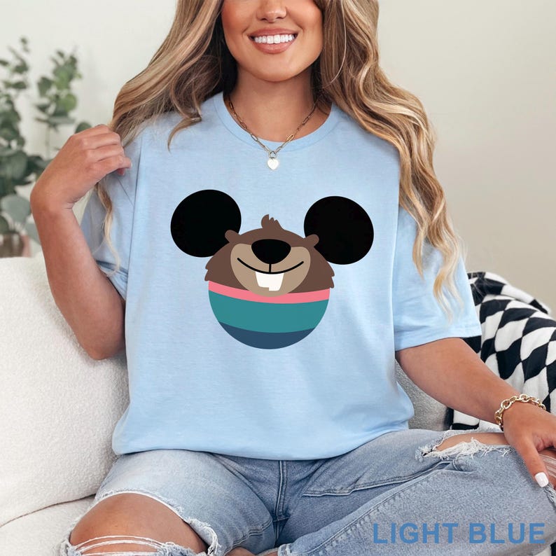 May include: Light blue t-shirt featuring a cartoon character design. The design includes a brown face with black ears, a pink stripe, and a teal and blue striped body. The text "LIGHT BLUE" is printed at the bottom.