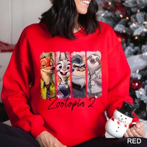 Puede incluir: Sudadera roja con personajes de Zootopia 2. El dise&ntilde;o incluye a Nick Wilde, Judy Hopps, el jefe Bogo y Flash. El texto "Zootopia 2" est&aacute; escrito en negro debajo de los personajes.