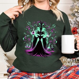 Op de afbeelding: Bosse groen sweatshirt met een afbeelding van een donkere tovenares met wervelende groene en paarse magie. De tovenares houdt drankflessen vast. De sweatshirt is effen van kleur met de woorden "FOREST GREEN" rechtsonder.