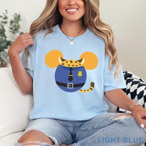 May include: Light blue t-shirt featuring a cartoon design of a police officer with mouse ears and a leopard print head. The design includes a blue uniform, a black tie, and a gold badge. The text "LIGHT BLUE" is at the bottom.