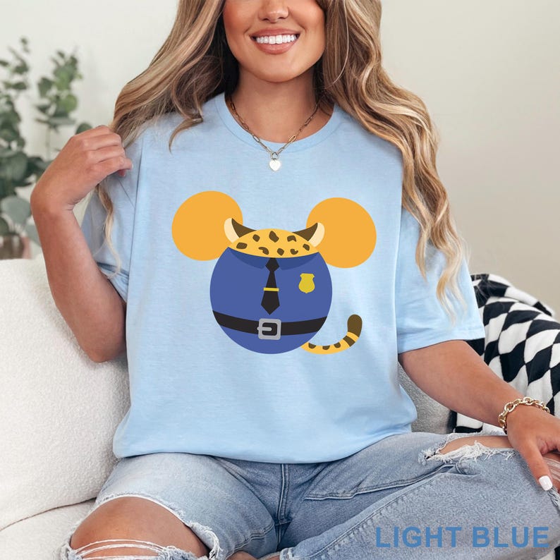 May include: Light blue t-shirt featuring a cartoon design of a police officer with mouse ears and a leopard print head. The design includes a blue uniform, a black tie, and a gold badge. The text "LIGHT BLUE" is at the bottom.