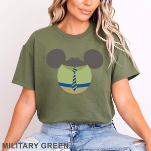 May include: Military green t-shirt featuring a cartoon character design. The design includes gray ears, a green face with a blue and yellow striped tie, and the text "MILITARY GREEN" at the bottom.