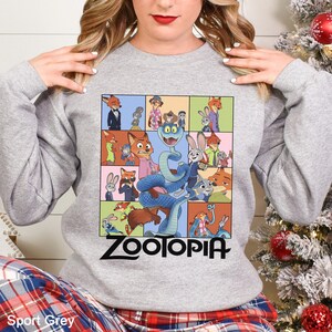 May include: Sport grey sweatshirt featuring a colorful Zootopia graphic. The graphic includes various characters from the animated film, such as Nick Wilde, Judy Hopps, and others, in a grid-like design. The word "Zootopia" is printed in black.