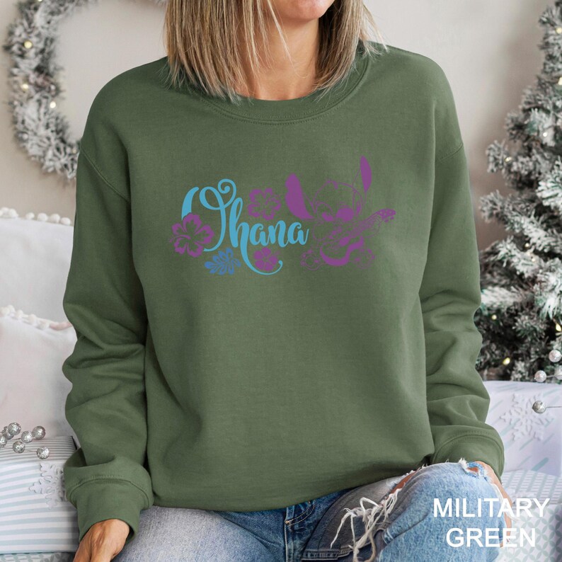 May include: Olive green sweatshirt with the word "Ohana" in blue and purple script, accompanied by floral and character graphics. The sweatshirt is a long-sleeved crew neck. The text "MILITARY GREEN" is printed in white at the bottom right.