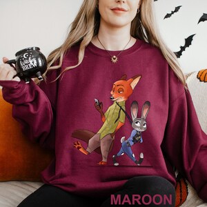 May include: Maroon sweatshirt featuring a cartoon fox and bunny character design. The fox is holding a phone, and the bunny is in a police uniform. The word "MAROON" is printed at the bottom. The sweatshirt is a crew neck style.