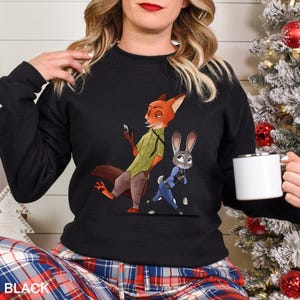 May include: Black crewneck sweatshirt featuring a cartoon image of Nick Wilde and Judy Hopps from Zootopia. Nick is depicted in a green shirt and Judy in a blue police uniform. The word "BLACK" is printed at the bottom.