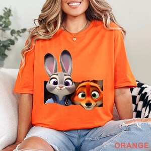 May include: Orange t-shirt featuring a graphic of Judy Hopps and Nick Wilde from Zootopia. The cartoon characters are in a rectangular frame. The word "ORANGE" is printed at the bottom of the shirt.