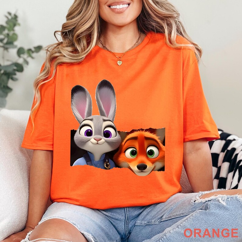 May include: Orange t-shirt featuring a graphic of Judy Hopps and Nick Wilde from Zootopia. The cartoon characters are in a rectangular frame. The word "ORANGE" is printed at the bottom of the shirt.