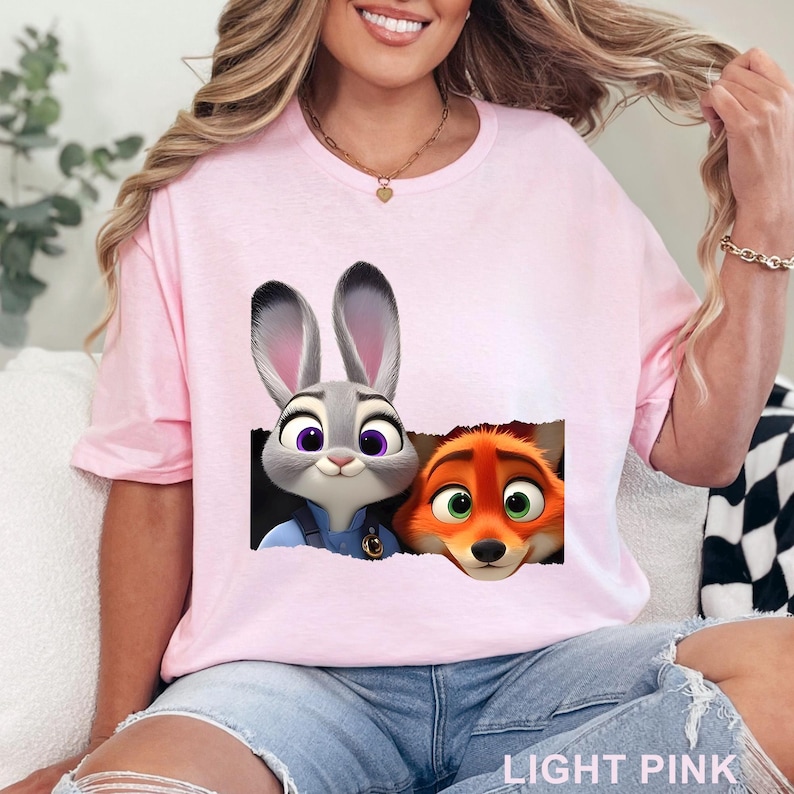 May include: Light pink t-shirt featuring a graphic of Judy Hopps and Nick Wilde from Zootopia. The characters are depicted in a torn paper effect. The text "LIGHT PINK" is printed at the bottom of the shirt.