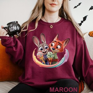 May include: Maroon sweatshirt featuring a colorful graphic of Judy Hopps and Nick Wilde from Zootopia holding ice cream cones. The word "MAROON" is printed at the bottom. A black mug with the text "Witches Brew" is held in the upper left corner.