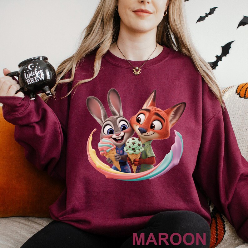 May include: Maroon sweatshirt featuring a colorful graphic of Judy Hopps and Nick Wilde from Zootopia holding ice cream cones. The word "MAROON" is printed at the bottom. A black mug with the text "Witches Brew" is held in the upper left corner.