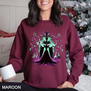 Op de afbeelding: Bordeaux sweatshirt met een afbeelding van een heksachtige figuur met groene en paarse accenten. Het ontwerp bevat drankflessen en wervelende rook. Het woord "MAROON" staat onderaan afgedrukt.