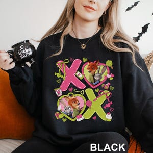 Peut inclure: Sweat-shirt noir avec un motif graphique repr&eacute;sentant Nick Wilde et Judy Hopps de Zootopie, dans des c&oelig;urs roses et verts. Le motif comprend les lettres X et O, des c&oelig;urs, des n&oelig;uds et une cl&eacute;. Le mot "BLACK" est imprim&eacute; en bas.