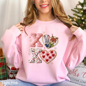 May include: Light pink sweatshirt with embroidered "XO" and heart designs. Features a heart with Judy Hopps and Nick Wilde from Zootopia. The sweatshirt has a crew neck and long sleeves. The text "Light Pink" is at the bottom left.