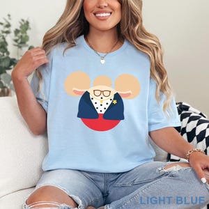 May include: Light blue t-shirt featuring a cartoon character design. The design includes a mouse-like face with glasses, a navy blue jacket, and a red circle base. The text "LIGHT BLUE" is at the bottom of the shirt.
