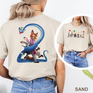 K&ouml;nnte beinhalten: Sandfarbenes T-Shirt mit einer farbenfrohen Grafik von Charakteren aus einem beliebten Animationsfilm. Auf der R&uuml;ckseite des Shirts ist eine blaue Schlange zu sehen, die sich um einen Fuchs, ein Kaninchen und eine Figur windet. Die Vorderseite hat eine kleinere Grafik von Charakteren und Herzen.
