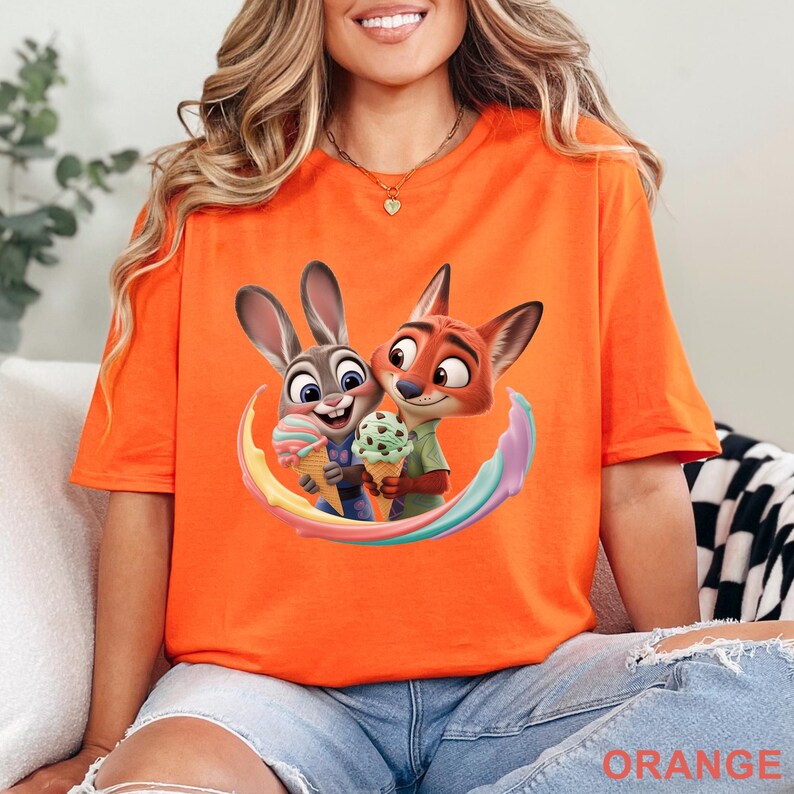May include: Orange t-shirt featuring a cartoon image of Judy Hopps and Nick Wilde from Zootopia, holding ice cream cones. The design includes a rainbow-colored arc. The word "ORANGE" is printed at the bottom of the shirt.