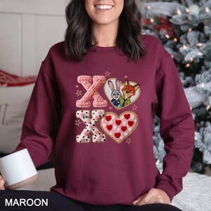 May include: Maroon sweatshirt with an embroidered design featuring the letters "XO" and images of a bunny and fox in heart-shaped frames. The "X" letters have a knitted and polka dot design, and the "O" has a heart pattern.