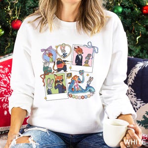 May include: White sweatshirt featuring a collage of cartoon characters in polaroid-style frames. The design includes various animal characters in different poses and settings, with colorful accents and the word "ZOOTOPIA".