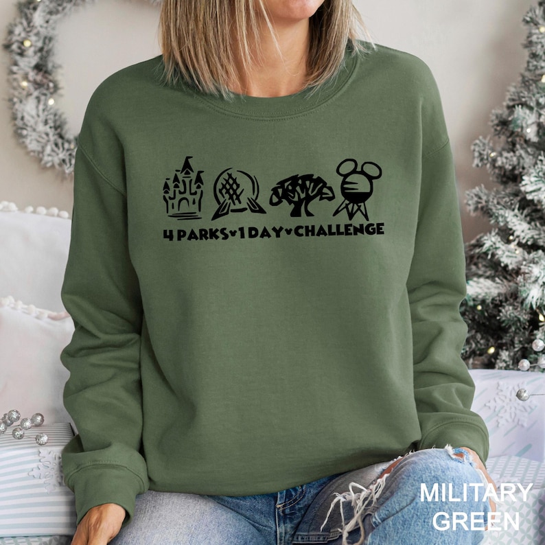 May include: Olive green sweatshirt with a black graphic of a castle, a seashell, a tree, and a Mickey Mouse head, with the text "4 PARKS 1 DAY CHALLENGE". The bottom right corner of the sweatshirt has the text "MILITARY GREEN".
