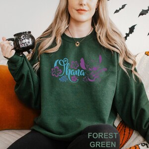 May include: Forest green sweatshirt with the word "Ohana" in blue and purple script, along with a purple cartoon character and floral accents. The sweatshirt also has the words "Forest Green" printed at the bottom.