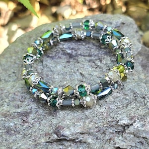 May include: A beaded bracelet with a double strand design. The bracelet features a mix of green, silver, and iridescent beads. The beads are various shapes and sizes, creating a textured look. The bracelet is resting on a gray stone surface.