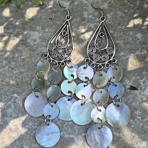 May include: Silver-toned dangle earrings with a teardrop-shaped top featuring filigree and a small crystal. Below, multiple strands of iridescent, round, shell-like discs hang down, creating a layered effect. The earrings are designed for pierced ears.