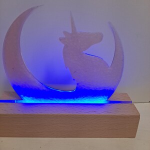 May include: A light-up unicorn silhouette inside a crescent moon, with a blue LED light strip at the base. The unicorn and moon are a translucent white, set on a rectangular wooden base.