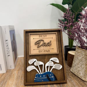 Peut inclure: Plaque en bois avec les mots "BEST Dad BY PAR" et un sac de golf avec des clubs. Les clubs de golf portent les noms de Jaxson, William et Grayson. La plaque est marron avec un fond marron clair.