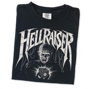 May include: A black t-shirt featuring a white graphic print of the character Pinhead from Hellraiser, with the text HELLRAISER in a distressed font above the character holding a puzzle box, surrounded by hanging chains and a Comfort Colors label.