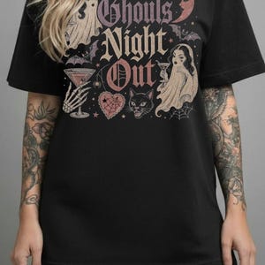Puede incluir: Una camiseta negra con un estampado gr&aacute;fico que incluye el texto Ghouls Night Out, dos figuras de fantasmas con copas de martini, un gato negro, murci&eacute;lagos, una luna creciente, una telara&ntilde;a y un coraz&oacute;n en tonos rosa, morado y crema.