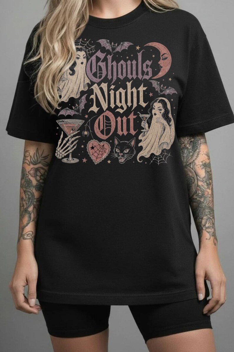 Puede incluir: Una camiseta negra con un estampado gr&aacute;fico que incluye el texto Ghouls Night Out, dos figuras de fantasmas con copas de martini, un gato negro, murci&eacute;lagos, una luna creciente, una telara&ntilde;a y un coraz&oacute;n en tonos rosa, morado y crema.