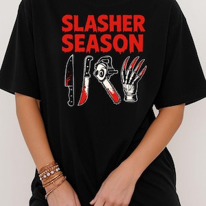 Könnte beinhalten: Schwarzes T-Shirt mit dem roten Text "SLASHER SEASON" über einer Grafik eines blutigen Messers, einer Kettensäge und eines Handschuhs mit langen roten Krallen. Das Shirt hat einen lässigen Stil.